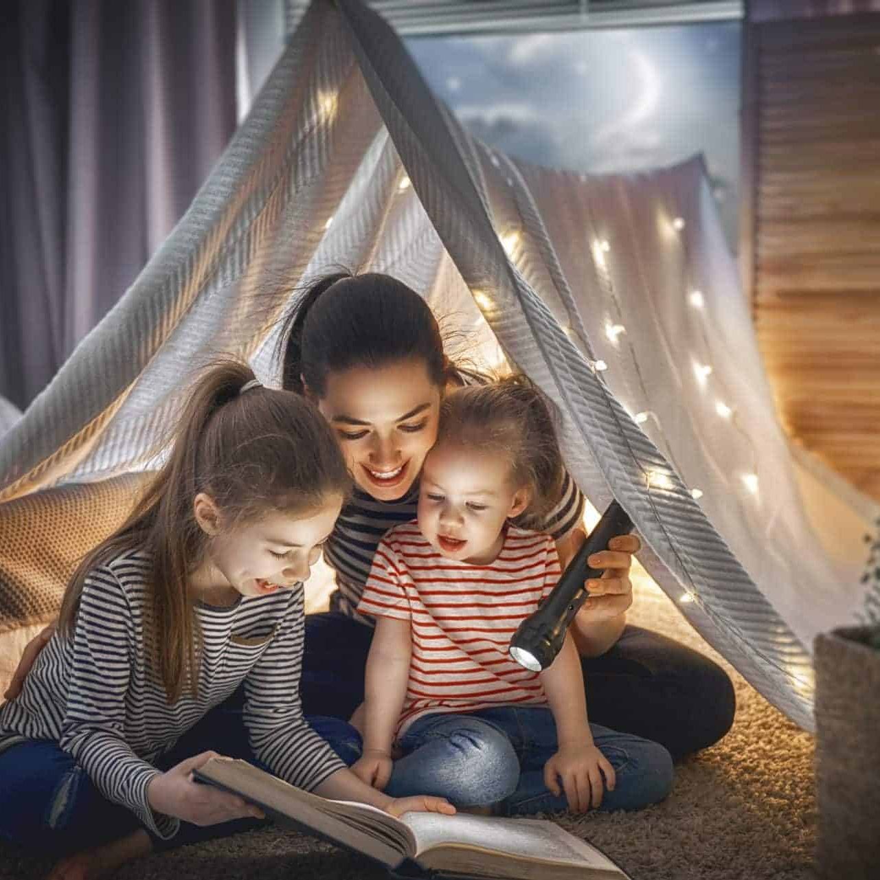Family bedtime. Mom and children daughters are reading a book in tent. Pretty young mother and lovely girls having fun in children room.